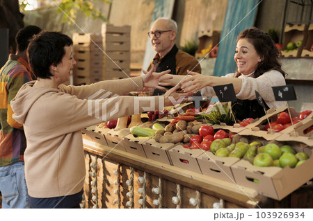 Happy local vendor greeting regular customer at food market stand with fresh organic produce, friendly farmer shaking hand of consumer. Selling homegrown food at farm market to happy clients. Happy local vendor greeting regular customer at food market stand with fresh organic produce, friendly farmer shaking hand of consumer. Selling homegrown food at farm market to happy clients. 103926934
