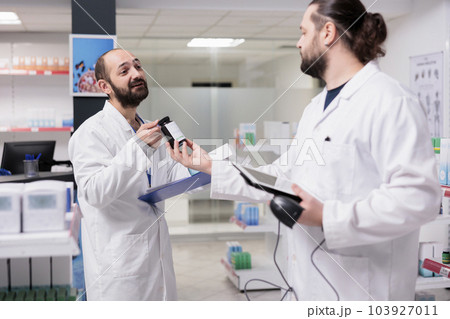 Drugstore employees looking at pills bottle checking medical label during pharmacy inventory, looking at clipboard full with drugs information. Pharmacists is responsible for organizing medication Drugstore employees looking at pills bottle checking medical label during pharmacy inventory, looking at clipboard full with drugs information. Pharmacists is responsible for organizing medication 103927011
