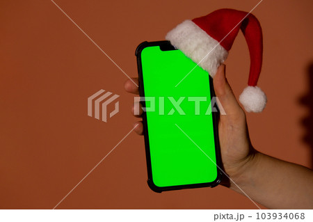 Mobile phone dressed in Santa-Claus red-white hat with chroma key screen against red background. Concept for Christmas or New Years holidays. Female Hand holding Blank cell phone. Digital gadget 103934068