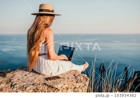 Digital nomad, Business woman working on laptop by the sea. Pretty lady typing on computer by the sea at sunset, makes a business transaction online from a distance. Freelance, remote work on vacation 103934362