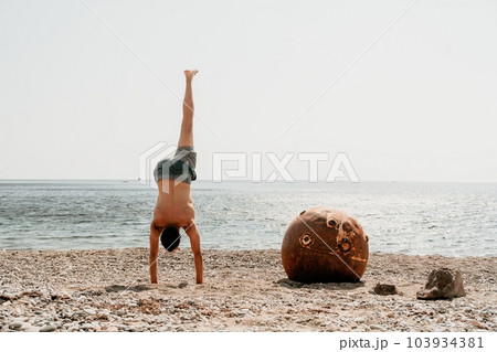 Man doing yoga exercise outdoors standing on old rusty floating marine mine on the beach with rocky shore and sea background. Healthy lifestyle, pollution, nature protection, war and peace concept. Man doing yoga exercise outdoors standing on old rusty floating marine mine on the beach with rocky shore and sea background. Healthy lifestyle, pollution, nature protection, war and peace concept. 103934381