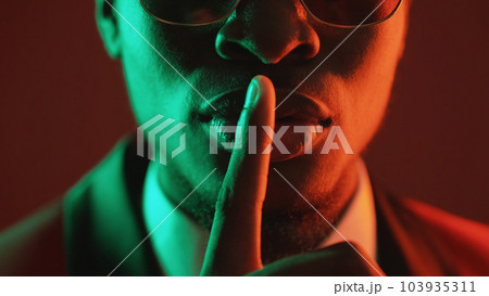 Shush gesture. Man secret. Confidential conspiracy. Green red neon light unrecognizable gentleman in suit glasses warning shh with finger on lips on dark. 103935311