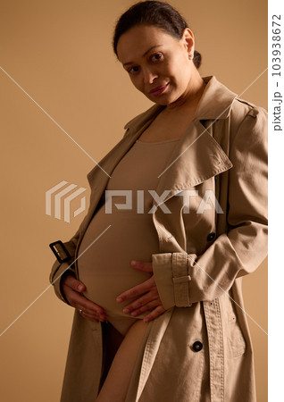Confident adult gorgeous pregnant woman in elegant trench coat under beige lingerie, strokes her belly, smiles looking at camera, isolated over studio cream background. Beautiful happy pregnancy Confident adult gorgeous pregnant woman in elegant trench coat under beige lingerie, strokes her belly, smiles looking at camera, isolated over studio cream background. Beautiful happy pregnancy 103938672