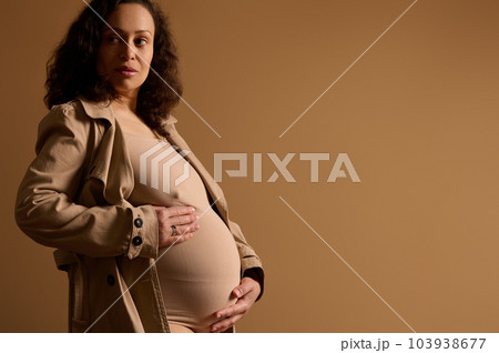 Confident adult gorgeous multi ethnic pregnant woman dressed in beige lingerie under trendy trench coat, strokes her belly, dreamily looking aside, poses isolated over cream background with copy space Confident adult gorgeous multi ethnic pregnant woman dressed in beige lingerie under trendy trench coat, strokes her belly, dreamily looking aside, poses isolated over cream background with copy space 103938677