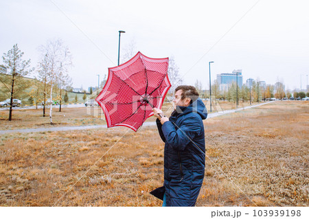 Shocked European white young male man fool around with umbrella in city park, strong storm wind, rain and bad weather. Wind broke umbrella. Autumn waist up lifestyle portrait. 103939198
