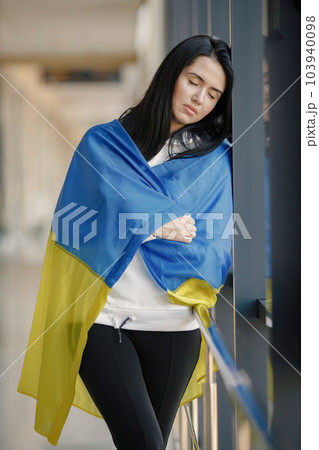 Woman with ukrainian flag posing for a photo. Young pretty girl standing in front of glass wall. Brunette woman wearing black clothes and support Ukraine. Woman with ukrainian flag posing for a photo. Young pretty girl standing in front of glass wall. Brunette woman wearing black clothes and support Ukraine. 103940098