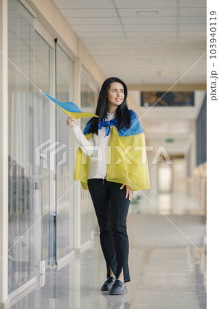 Woman with ukrainian flag posing for a photo. Young pretty girl standing in front of glass wall. Brunette woman wearing black clothes and support Ukraine. 103940119