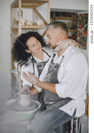 Mutual creative work. Adult elegant couple in casual clothes and aprons. People creating a bowl on a pottery wheel in a clay studio. 103940434