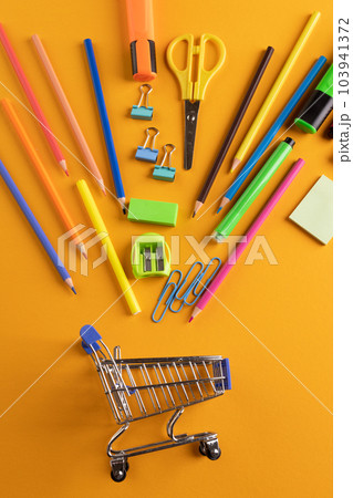 Close up of miniature shopping trolley with school materials on orange background Close up of miniature shopping trolley with school materials on orange background 103941372