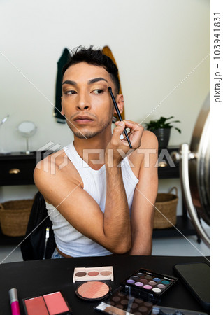 Biracial transgender man looking in mirror and putting on make-up, applying eyeshadow 103941831