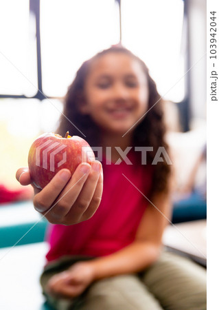 Smiling elementary schoolgirl showing apple during lunch break while sitting in school 103942044