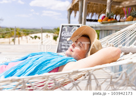 Caucasian senior woman wearing hat sleeping on hammock at beach during sunny day 103942173
