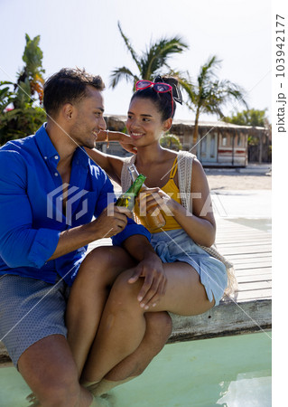 Caucasian smiling young couple toasting beer bottles while sitting at poolside at tourist resort 103942177