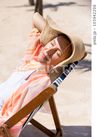 Caucasian senior woman wearing hat and sunbathing while sitting on deck chair at beach 103942208