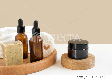 Glass bottles on wooden plate. Natural skin care SPA beauty product design. Mineral organic oil cosmetics on beige background. Earth tones. Mock-Up. Oily pipette. Face and body treatment. Front View 103943578