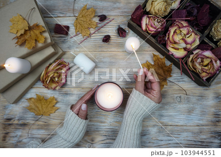 A woman lights an aromatic candle. There are dried roses in a box on the table and yellow maple leaves are lying. A woman lights an aromatic candle. There are dried roses in a box on the table and yellow maple leaves are lying. 103944551
