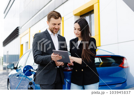 Smiling woman standing at the car and pointing at car seller tablet. She is picked right car for her she saw online. A woman concludes an agreement to buy a car and shakes the manager's hand 103945837