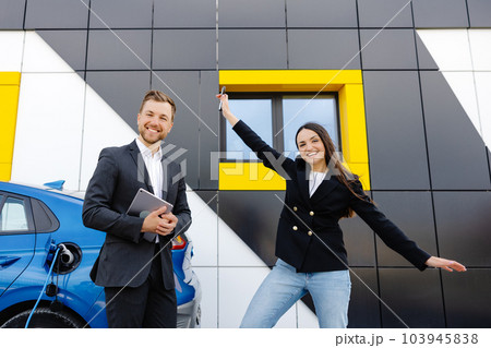 Happy female client standing at brand new car and taking keys from dealer in dealership outdoor. The woman is very happy with the new car, holding the keys in her hands 103945838