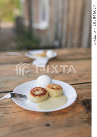 Two white plates with cheesecakes, sweet sauce, eggs and metal fork on aged wooden table. Village atmosphere and early morning breakfast for two persons, with old village house background. 103946027