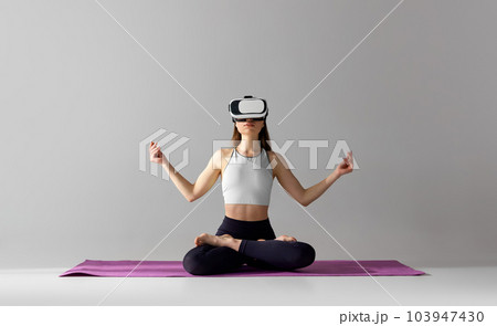 Young girl with slim body, wearing VR glasses and training in sportswear against grey studio background. Yoga and meditation session Young girl with slim body, wearing VR glasses and training in sportswear against grey studio background. Yoga and meditation session 103947430