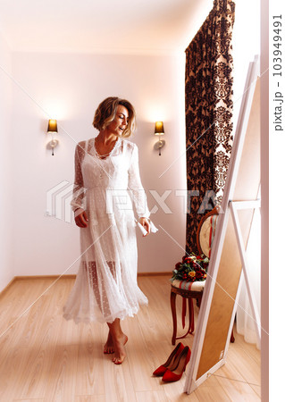 Bride in elegant white dress looking in mirror. The bride smiles and poses in front of the mirror. Bride in elegant white dress looking in mirror. The bride smiles and poses in front of the mirror. 103949491
