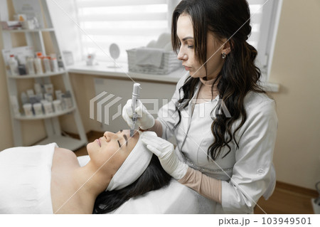 A professional cosmetologist conducts fractional mesotherapy to stimulate the natural process of facial skin restoration in a young woman in a beauty salon at a reception with a cosmetologist 103949501