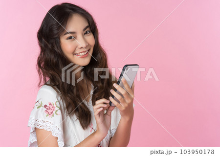 Young Asian woman texting message using mobile phone application looking directly at the camera in a studio shot on a pink background 103950178