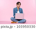 Young Asian college student sits cross-legged on floor working with a laptop computer, looking directly at the camera in a studio shot on a pink background 103950330