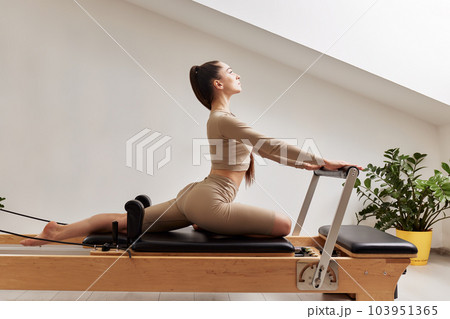 A woman is doing Pilates on a reformer bed in a bright studio. A