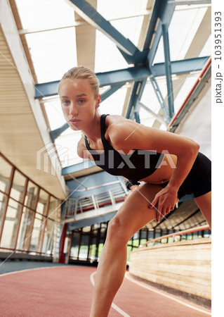 Young athlete girl is warming up before the race at the stadium. A slender blonde in a black tracksuit stretches her leg muscles before training. Sports and recreation. Young athlete girl is warming up before the race at the stadium. A slender blonde in a black tracksuit stretches her leg muscles before training. Sports and recreation. 103951393