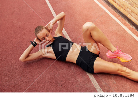 Young athlete girl is warming up before the race at the stadium. A slender blonde in a black tracksuit stretches her leg muscles before training. Sports and recreation. Young athlete girl is warming up before the race at the stadium. A slender blonde in a black tracksuit stretches her leg muscles before training. Sports and recreation. 103951394