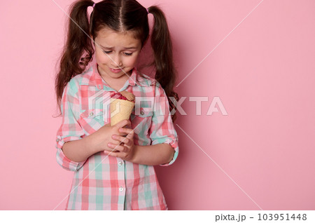 Adorable happy little child girl with two ponytails, wearing checkered shirt, holding a waffle cone with delicious ice cream over isolated pink background. Food. Summer 103951448