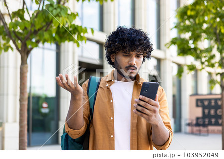 Upset student with backpack reads bad news on phone, man with phone browses internet page on smartphone, opens app, hispanic outside near university campus. 103952049