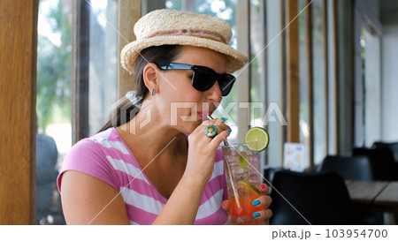 Young woman in straw hat and sunglasses drinking refreshing lemonade in cafe at hot summer day 103954700