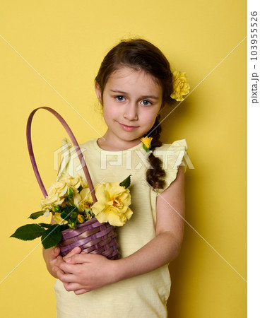 Beauty portrait Caucasian noble charming little girl in yellow elegant wear and flowers in hair, holding bouquet of yellow roses in purple wicker basket, smiling looking at camera, isolated background Beauty portrait Caucasian noble charming little girl in yellow elegant wear and flowers in hair, holding bouquet of yellow roses in purple wicker basket, smiling looking at camera, isolated background 103955526