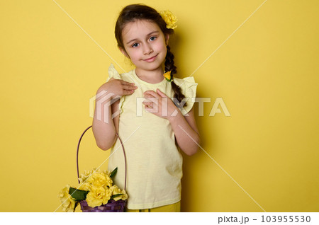 Emotional portrait of an adorable little child girl wearing yellow dress, holding purple wicker basket with yellow roses, smiling cutely looking at camera, isolated over yellow color studio background Emotional portrait of an adorable little child girl wearing yellow dress, holding purple wicker basket with yellow roses, smiling cutely looking at camera, isolated over yellow color studio background 103955530