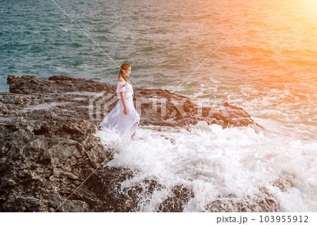 A woman stands on a rock in the sea during a storm. Dressed in a A woman stands on a rock in the sea during a storm. Dressed in a 103955912