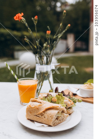Delicious baguette sandwich with Fresh orange juice on marble table on the cafe terrace on the street. Selective focus. Delicious baguette sandwich with Fresh orange juice on marble table on the cafe terrace on the street. Selective focus. 103956074