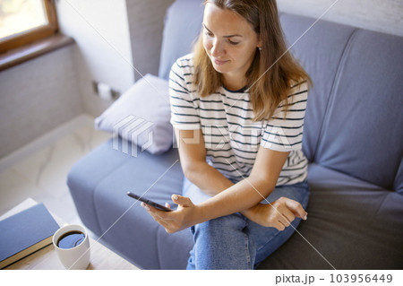 Woman sitting on sofa in living room, browsing wireless internet on smartphone Woman sitting on sofa in living room, browsing wireless internet on smartphone 103956449