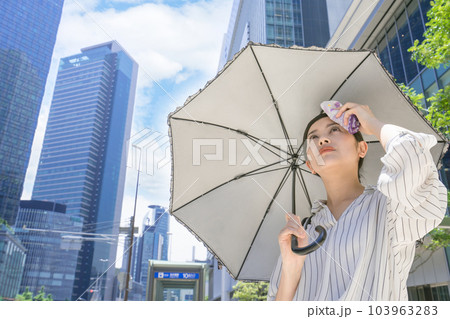 高層ビル街で日傘をさして、タオルで汗を拭うビジネスウーマン 高層ビル街で日傘をさして、タオルで汗を拭うビジネスウーマン 103963283