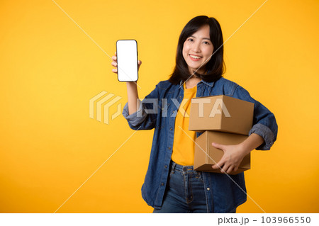 Portrait asian young woman wearing yellow t-shirt and denim shirt holding mobile phone and parcel box isolated on yellow studio background, Delivery courier and shipping service concept. 103966550
