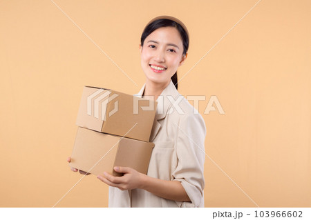 portrait of happy successful confident young asian business woman wearing white jacket holding parcel box isolated on beige studio background, Delivery courier and shipping service concept. portrait of happy successful confident young asian business woman wearing white jacket holding parcel box isolated on beige studio background, Delivery courier and shipping service concept. 103966602