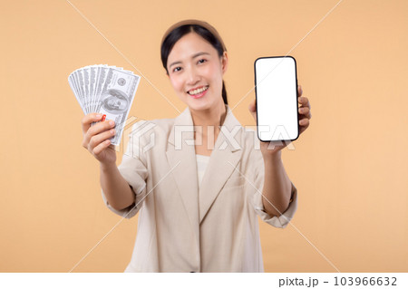 portrait of happy successful confident young asian business woman wearing white jacket holding smartphone and cash money dollar standing over beige background. millionaire business, shopping concept. portrait of happy successful confident young asian business woman wearing white jacket holding smartphone and cash money dollar standing over beige background. millionaire business, shopping concept. 103966632