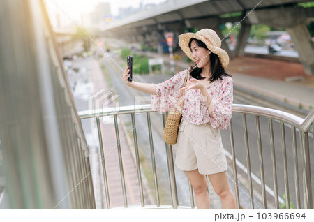 young asian woman traveler with weaving basket using mobile phone and standing on overpass with railway background. Journey trip lifestyle, world travel explorer or Asia summer tourism concept. 103966694