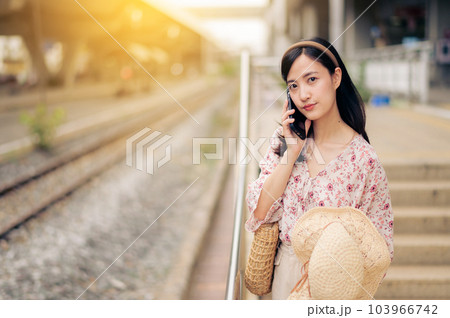 Asian young woman traveler with weaving hat using a mobile phone beside railway train station in Bangkok. Journey trip lifestyle, world travel explorer or Asia summer tourism concept. 103966742