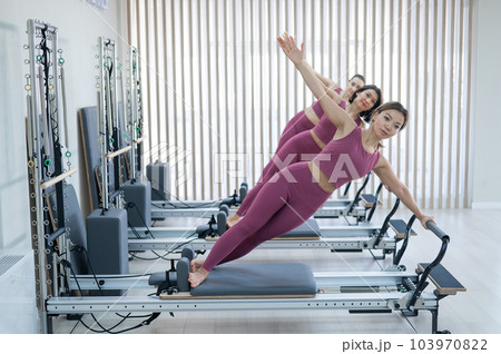 Three Asian women in pink sportswear doing side planks on a reformer. Pilates classes. Three Asian women in pink sportswear doing side planks on a reformer. Pilates classes. 103970822