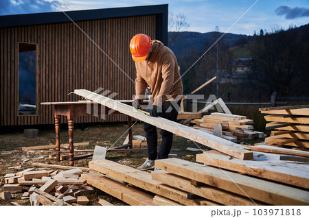 Man worker building wooden frame house. Carpenter using tape measure for measuring wooden planks and making marks with pencil. Carpentry concept. 103971818