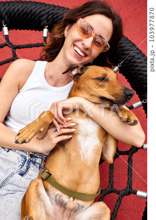 Charming young smiling girl is resting on a swing in the city yard with a golden dog. The girl hugs her pet. Love and affection between owner and pet. Adopting a pet from a shelter. Close-up. 103977870