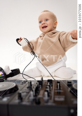 A cute smiling little child in a beige hoodie is sitting on the floor with dj headphones and a dj mixing board. Music and fun. Isolated on white background. A cute smiling little child in a beige hoodie is sitting on the floor with dj headphones and a dj mixing board. Music and fun. Isolated on white background. 103977886