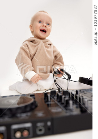 A cute smiling little child in a beige hoodie is sitting on the floor with dj headphones and a dj mixing board. Music and fun. Isolated on white background. A cute smiling little child in a beige hoodie is sitting on the floor with dj headphones and a dj mixing board. Music and fun. Isolated on white background. 103977887